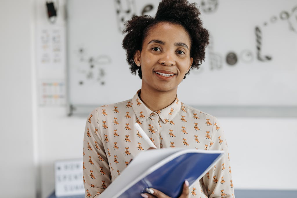 confident female teacher smiling while holding a notebook in a classroom setting. 8423065